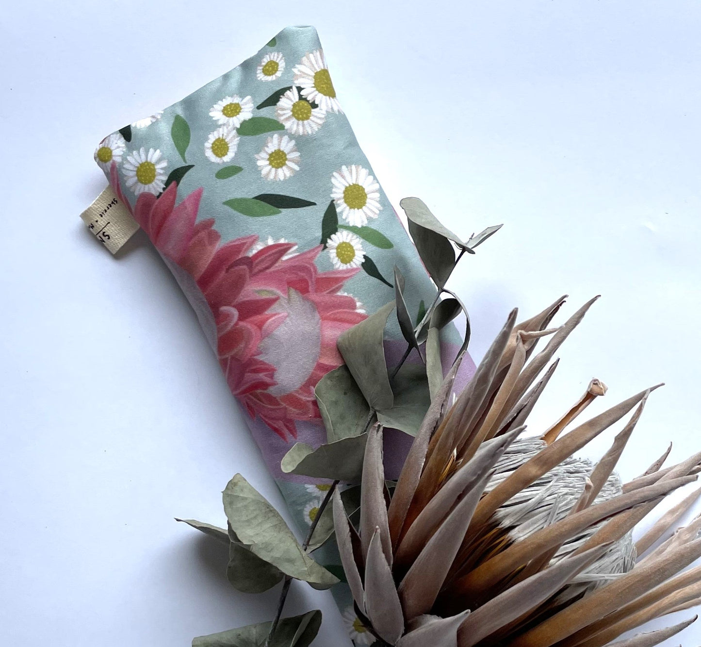 Sherrie Morgan Design relaxing eye pillow with a soft protea and daisy flower print in pink and turquoise, pictured with a dried king protea flower