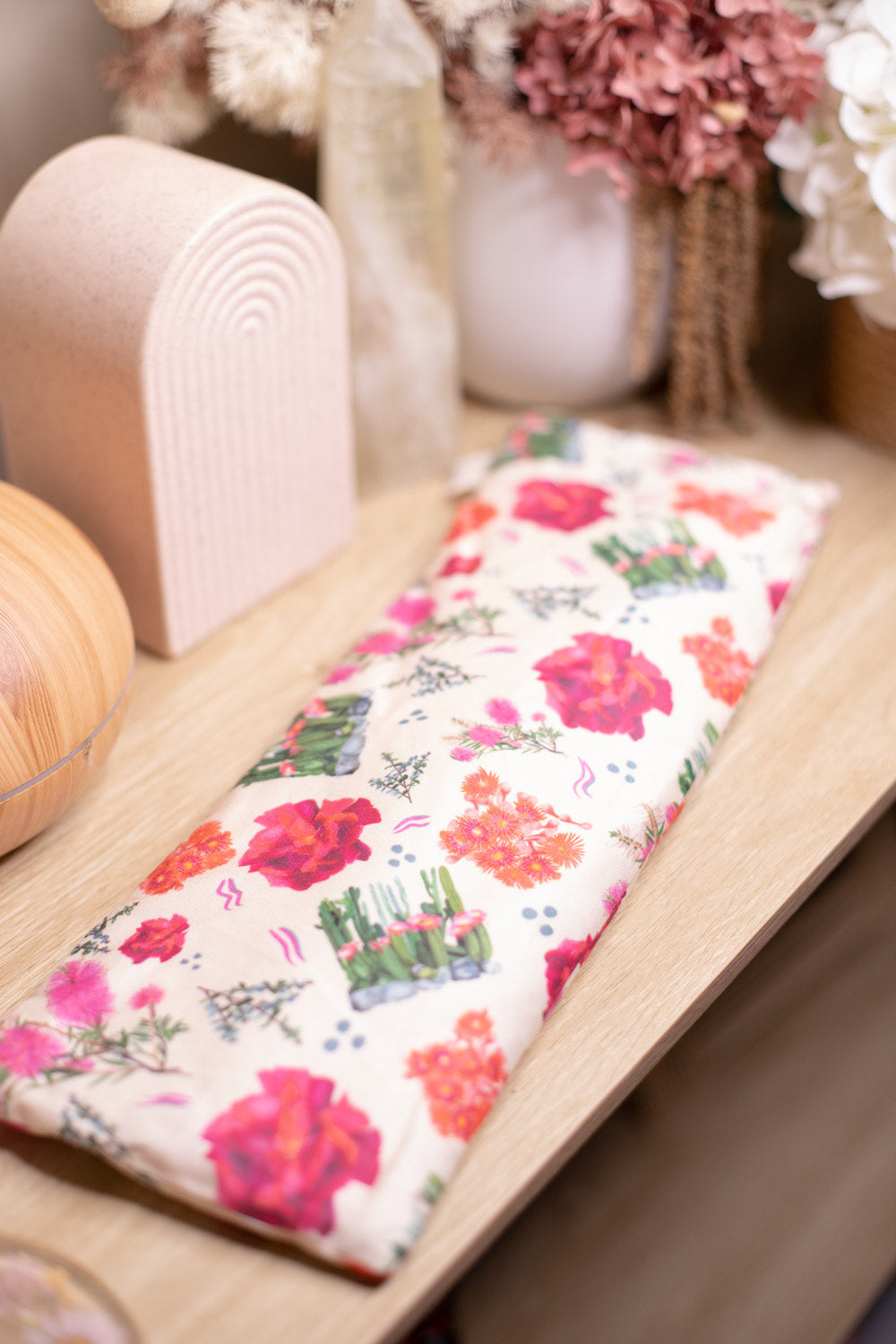 Sherrie Morgan Design relaxing wheat, heat pillow with a cactus, rose and bottlebrush flower print in pink, red and cream, displayed with a vase.