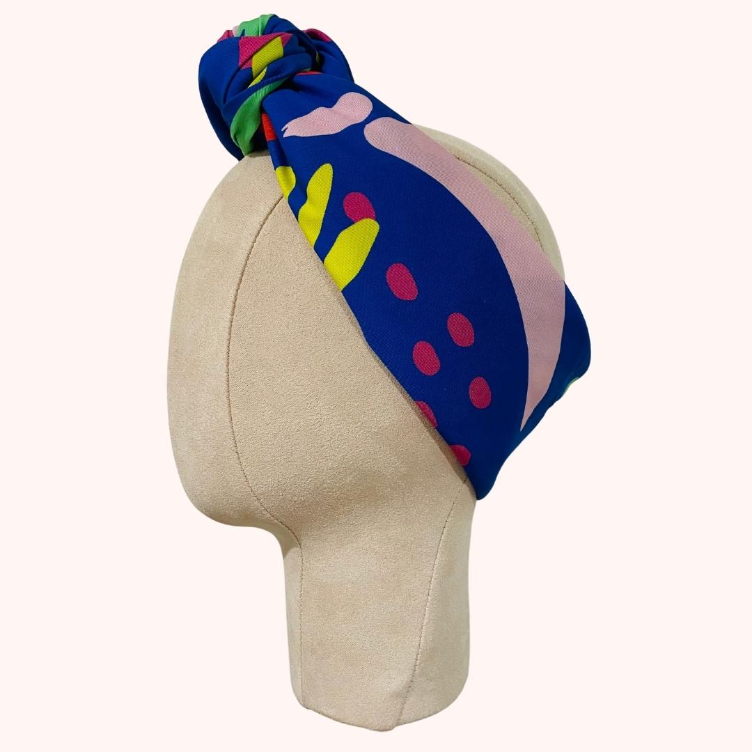 SMD scarf, Nerida Hansen print featuring large red, yellow and green sunshine and flowers on a blue base, displayed as a headband with top knot, side view