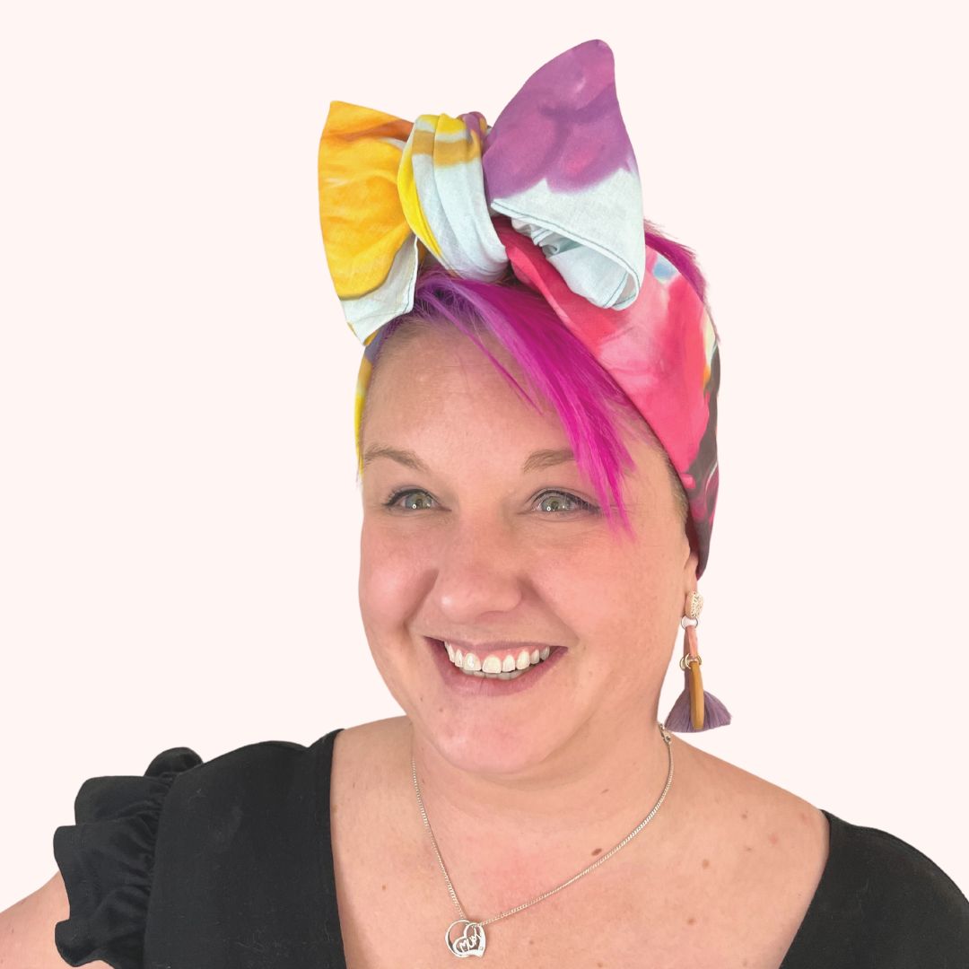 SMD original 'Floral Frenzy', features multi coloured flowers including protea, lavneder and billy button on a light blue base, scarf displayed as headwrap with bow, front view