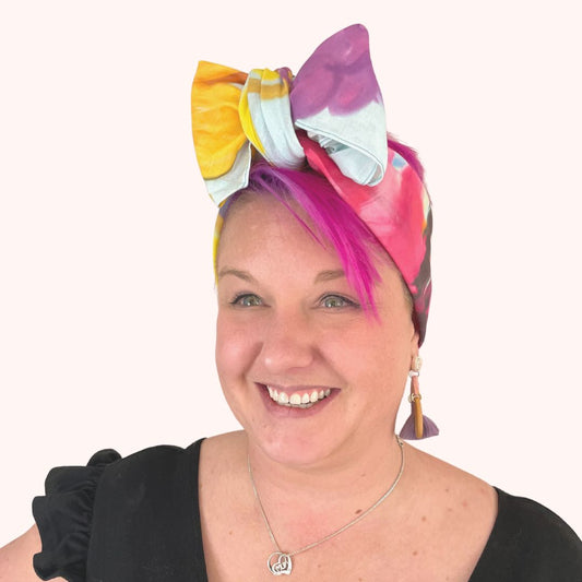 SMD original 'Floral Frenzy', features multi coloured flowers including protea, lavneder and billy button on a light blue base, scarf displayed as headwrap with bow, front view