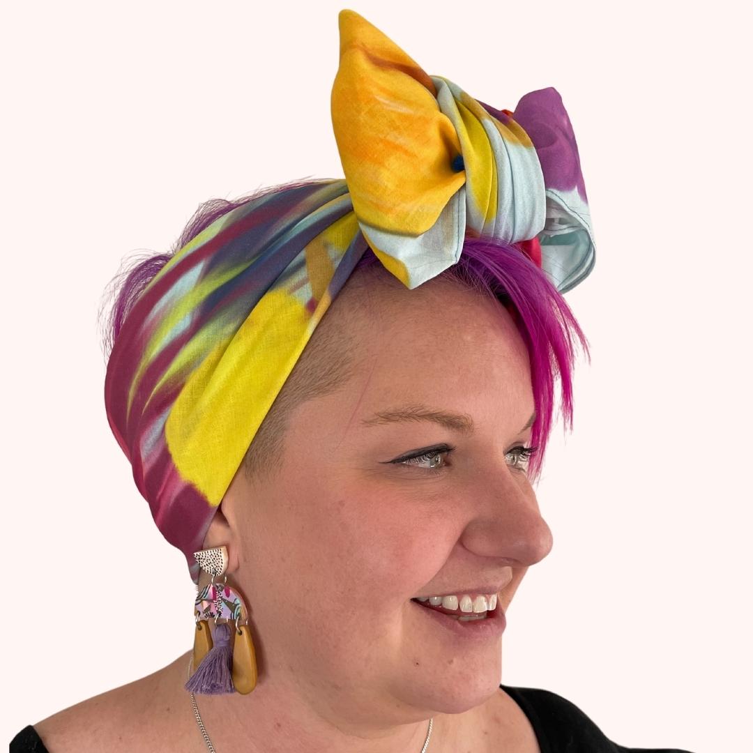 SMD original 'Floral Frenzy', features multi coloured flowers including protea, lavneder and billy button on a light blue base, scarf displayed as headwrap with bow, side view