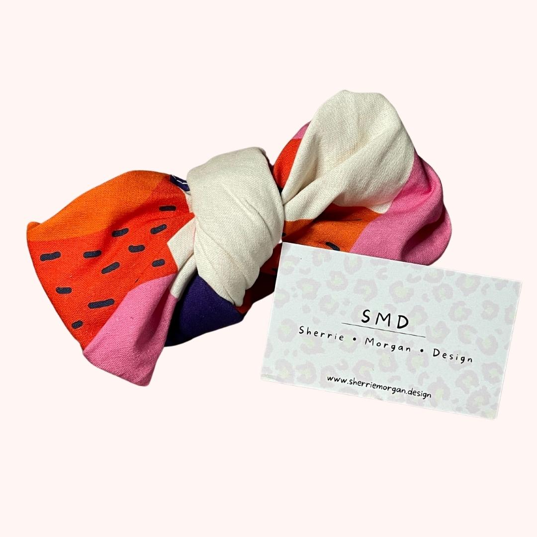 SMD scarf with Jennifer Bouron design of large shapes in purple, pink, orange and off white, displayed as a bow with business card