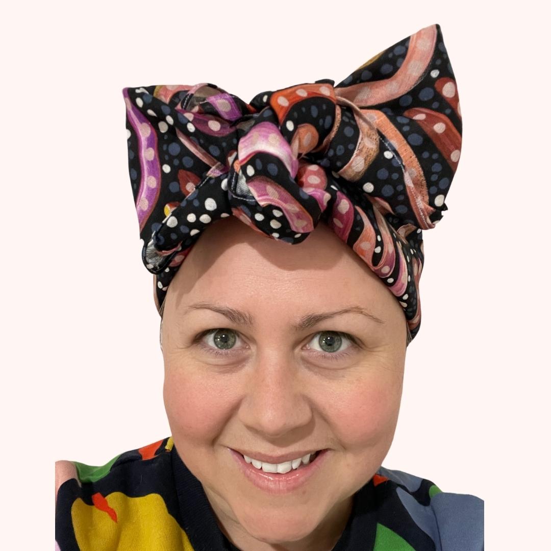 SMD scarf, charcoal base with pink, orange, white sandhills design by Holly Sanders, shown as a headscarf with large bow