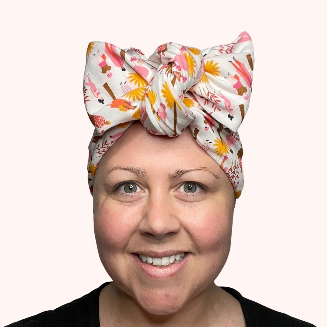 SMD scarf, white base with ladies and dogs walking in pink and yellow, shown as headband with a large bow