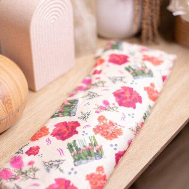 Sherrie Morgan Design relaxing wheat, heat pillow with a cactus, rose and bottlebrush flower print in pink, red and cream, displayed with a vase.