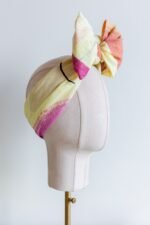 Original Sherrie Morgan design 'Desert Rainbow', comprises a yellow setting with rainbows and shapes in brown, pink and orange, displayed as a headscarf with a large bow, side view